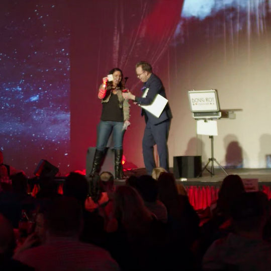 Girl on stage next to Doug. She is holding a card, waiting to find out if he guesses correctly.
